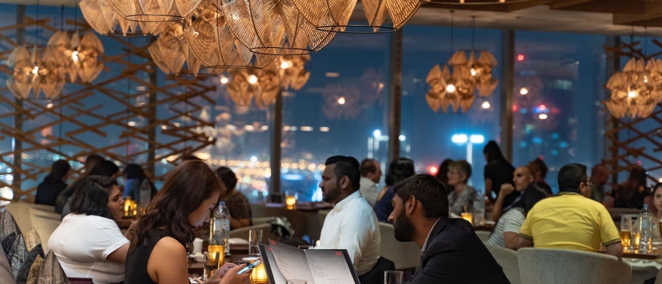 Diners at tables in a restaurant at night, under woven pendant lamps, with a lit city skyline through the windows behind them.